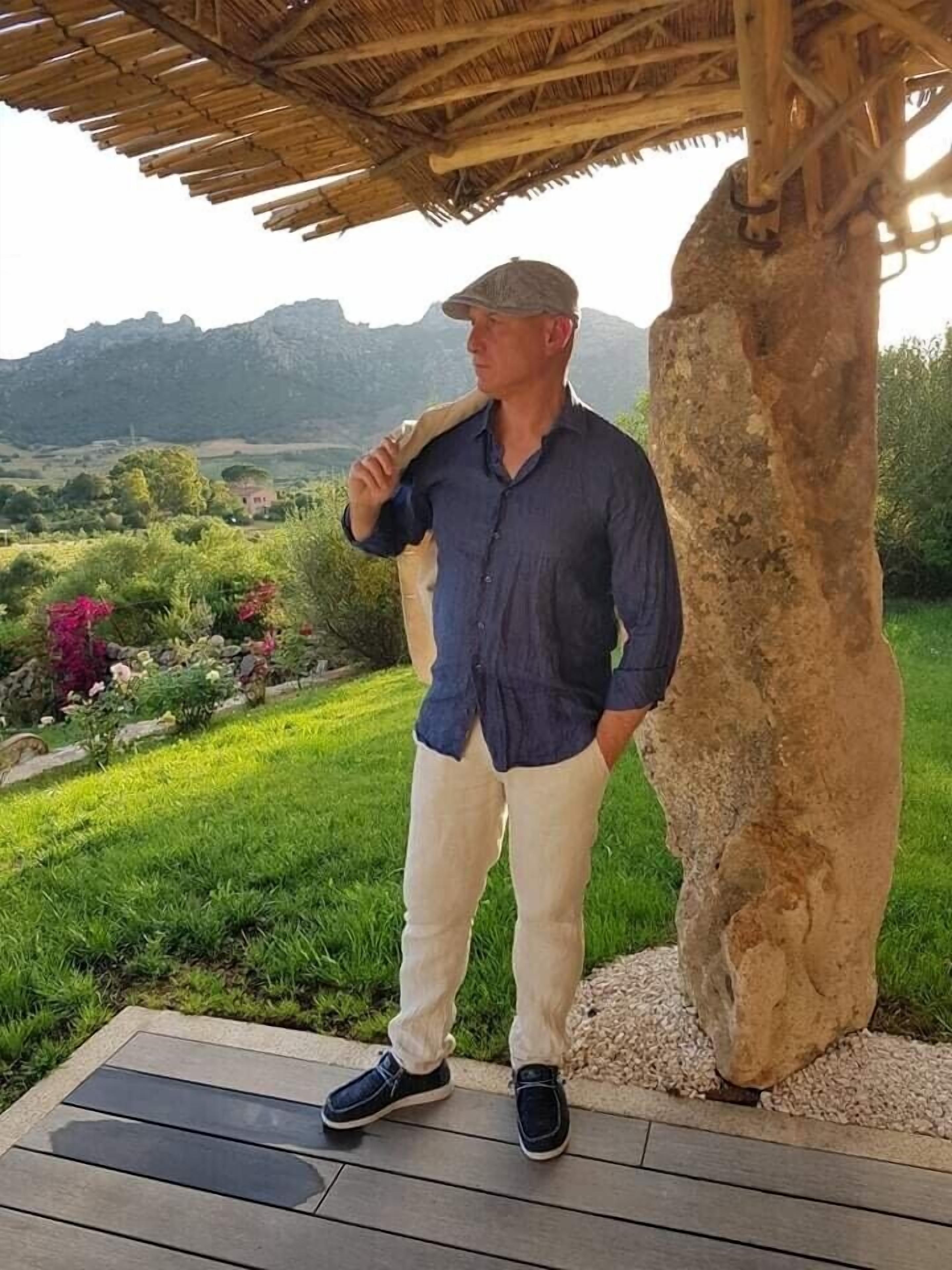 Un uomo in piedi sotto un pergolato di legno con un pilastro in pietra, che si affaccia su un paesaggio verde e rigoglioso con montagne sullo sfondo.