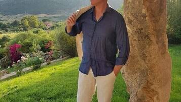 Un uomo in piedi sotto un pergolato di legno con un pilastro in pietra, che si affaccia su un paesaggio verde e rigoglioso con montagne sullo sfondo.