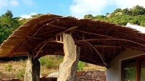 Una cabaña rústica con base de piedra, techo de paja y un muro de piedra que rodea la propiedad.