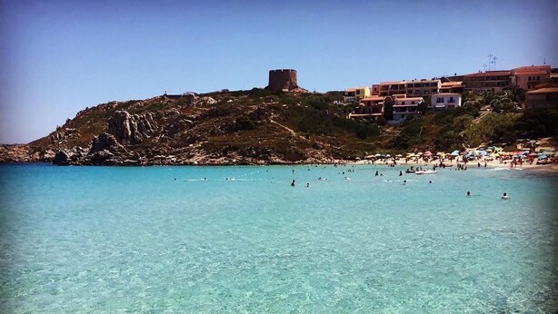 Una playa costera con agua turquesa cristalina, un litoral rocoso y una colina con edificios al fondo.