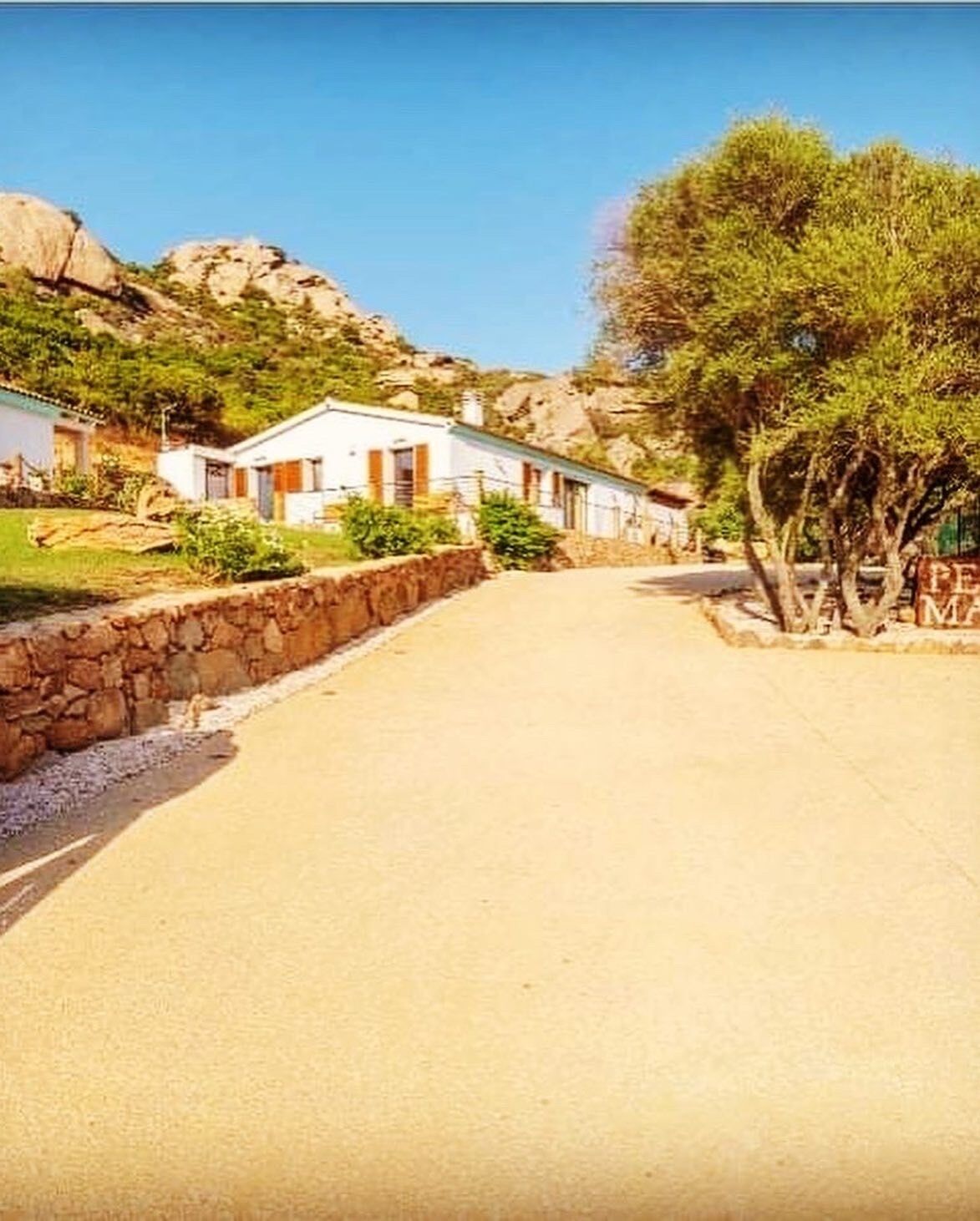 A road leading to a row of white buildings with stone walls, surrounded by greenery and hills.