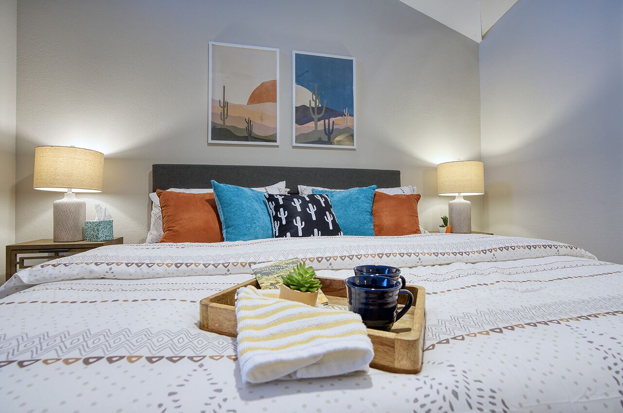 A neatly made bed with patterned bedding, two wall-mounted paintings, bedside lamps, a tray with a towel, a small plant, and a mug.