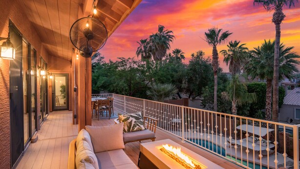 A wooden deck with a sofa and a fire feature, overlooking a swimming pool and palm trees under a sunset sky. - Amazing Peaceful Resort - Heated Pool, Hot Tub (Fountain Hills)