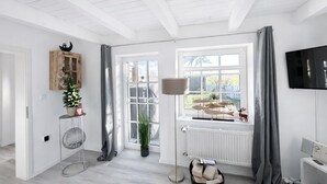 Een lichte kamer met een groot raam, een radiator, een televisie en een houten kast met decoratieve spullen.