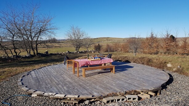 Een rond houten terras met een picknicktafel en bankjes, omringd door een stenen muur en een uitzicht op een landelijk landschap.