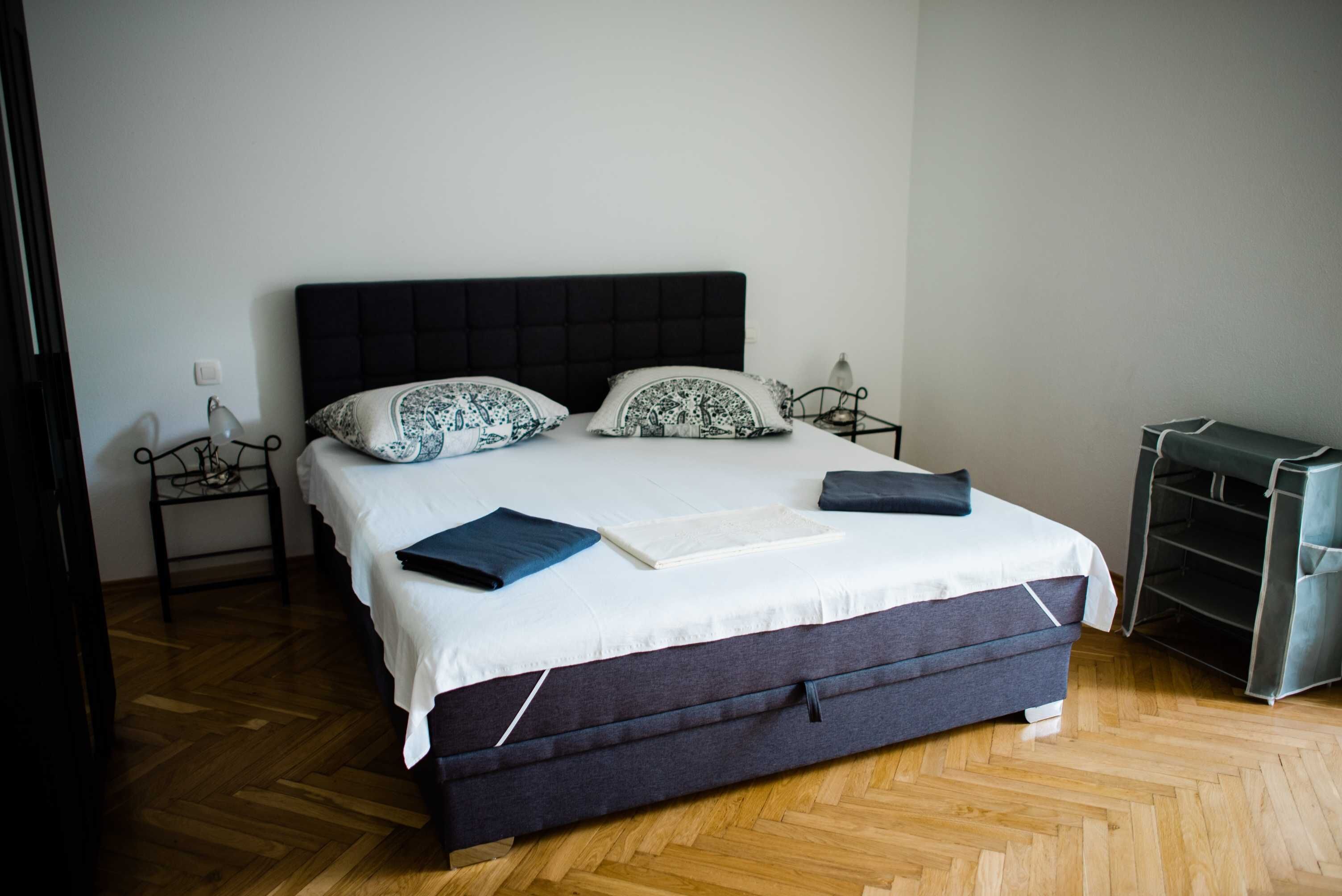 Un lit bien fait, avec une tête de lit sombre, une literie blanche et des coussins à motifs. On y trouve une table de chevet avec une lampe et une petite étag ère. La pièce est dotée d’un plancher en bois et d’une étagère métallique sur le côté droit.