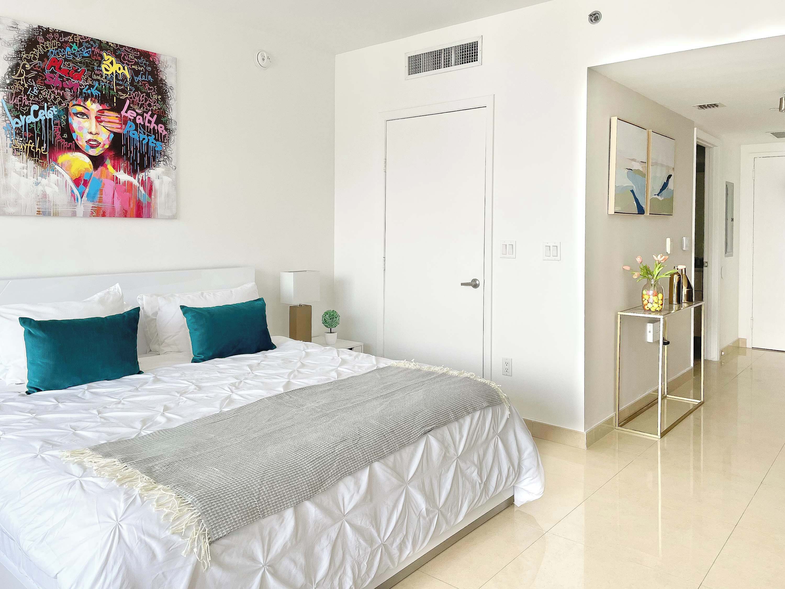 Een moderne slaapkamer met een groot bed, een nachtkastje met een lamp, een kleine potplant en een deur die leidt naar een andere kamer.