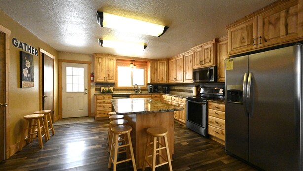 A kitchen with wooden cabinets, a stainless steel refrigerator, a large island with stools, and a window providing natural light. - Mountain View Cabin 22 Miles From Yellowstone (Island Park)