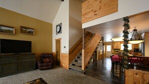 A spacious living area with a large flat-screen TV, a leather armchair, and a patterned rug. A wooden staircase leads to a dining area with red tablecloths and wooden chairs. - Mountain View Cabin 22 Miles From Yellowstone (Island Park)