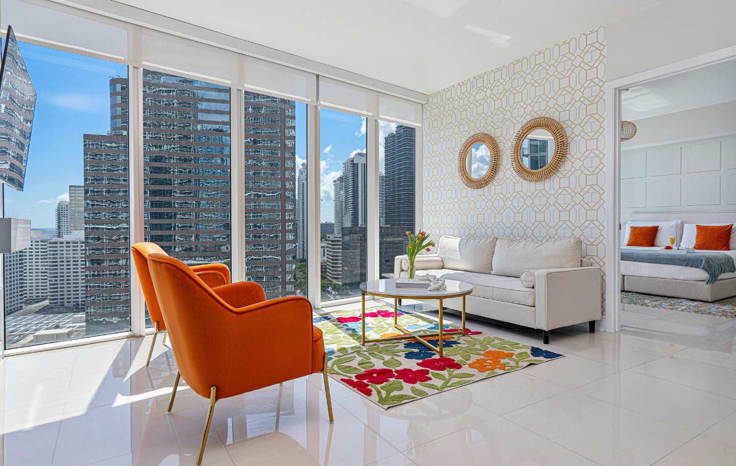 A modern living room with a city view, a white sofa, orange armchairs, and a patterned rug.