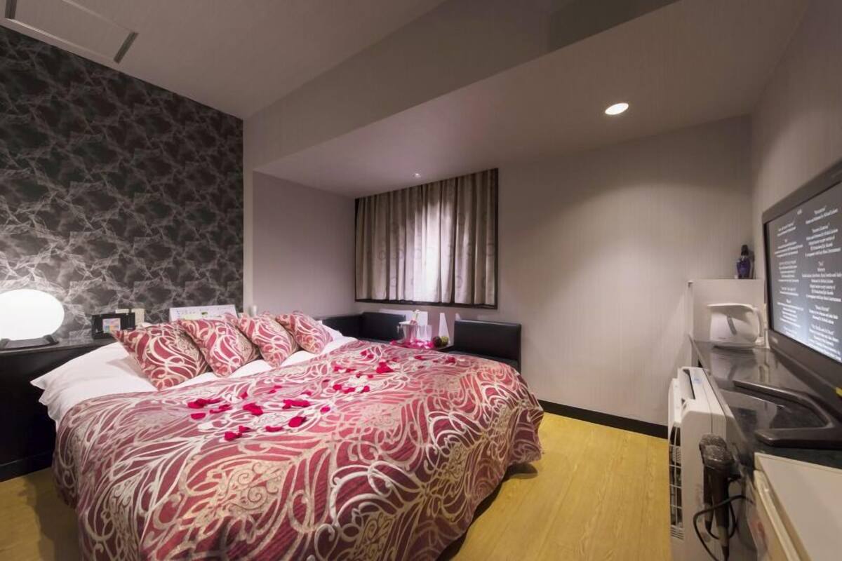 Una habitación de hotel con cama, escritorio, silla, lámpara y ventana con cortinas.