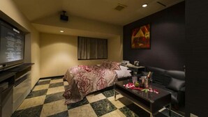 Habitación de hotel con piso a cuadros, cama con colcha floral, sofá de cuero negro, mesita con una botella y vasos, y televisor a la izquierda.