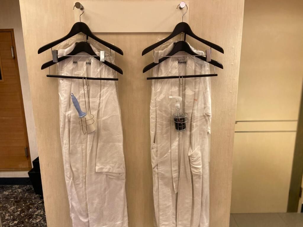 Dos batas blancas colgadas en perchas negras en una habitación con una puerta de madera y suelo de baldosas.