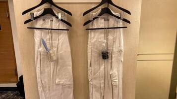 Dos batas blancas colgadas en perchas negras en una habitación con una puerta de madera y suelo de baldosas.