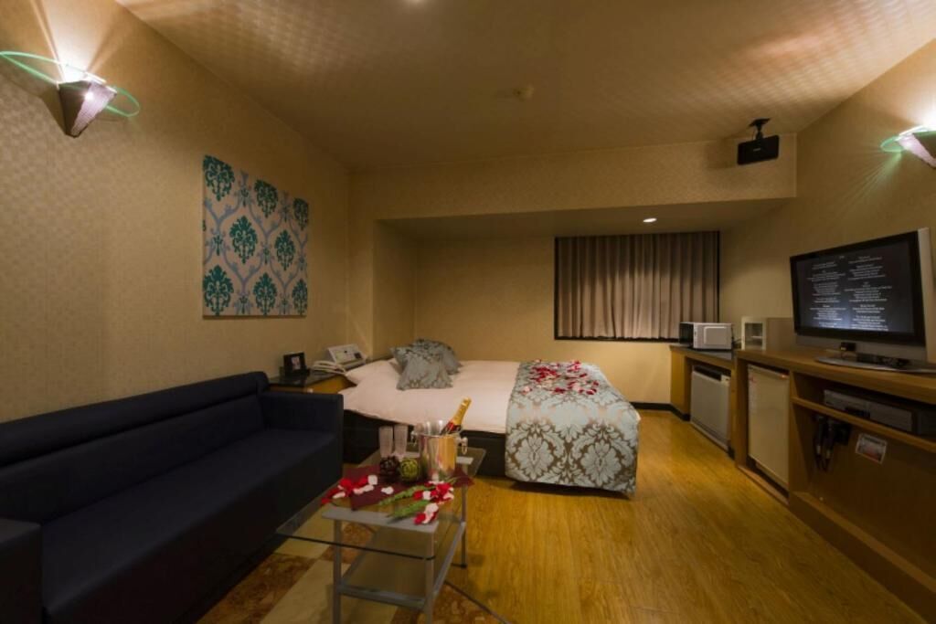 A hotel room with a bed, a sofa, a small table with a drink, a TV, and a projector on the ceiling.