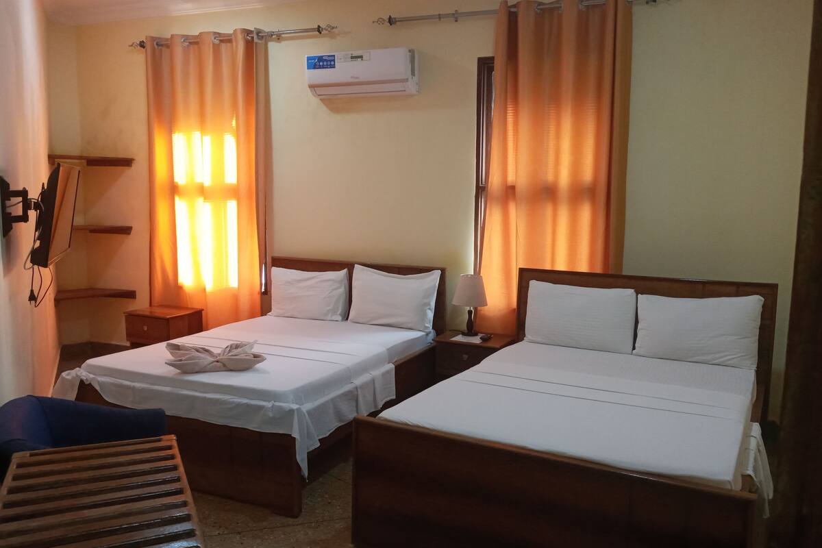 Una habitación de hotel con dos camas individuales, un banco de madera, una mesita de noche con lámpara y un equipo de aire acondicionado en la pared.