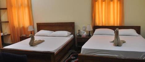Habitación de hotel con dos camas individuales, cada una con cabecero de madera, ropa blanca y un peluche sobre la cama. Hay una mesita de noche con lámpara, una mesita auxiliar con lámpara y un reloj de pared. La habitación tiene cortinas naranjas y una alfombra oscura en el suelo.