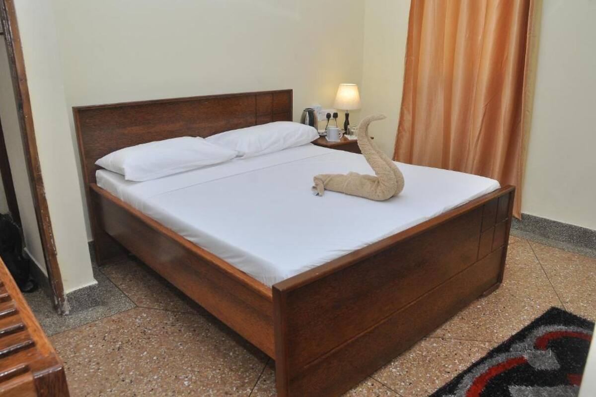 Una cama bien hecha con sábanas blancas, cabecera de madera, mesita de noche con lámpara y una figurita decorativa.