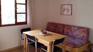 A room with a wooden table, two chairs, a colorful sofa, a window with curtains, an air conditioning unit, and a framed picture on the wall. - Exclusive Cottages in a Quiet Oliverove Near the sea (Kantanos-Selino)