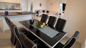 A modern kitchen with white cabinets, a dining area with a glass table and black chairs, and a living room with a sofa and TV. - Oceanview Deluxe Penthouse Apartment With 89m2 Living Space & 90m2 Roof Terrace (Okrug)
