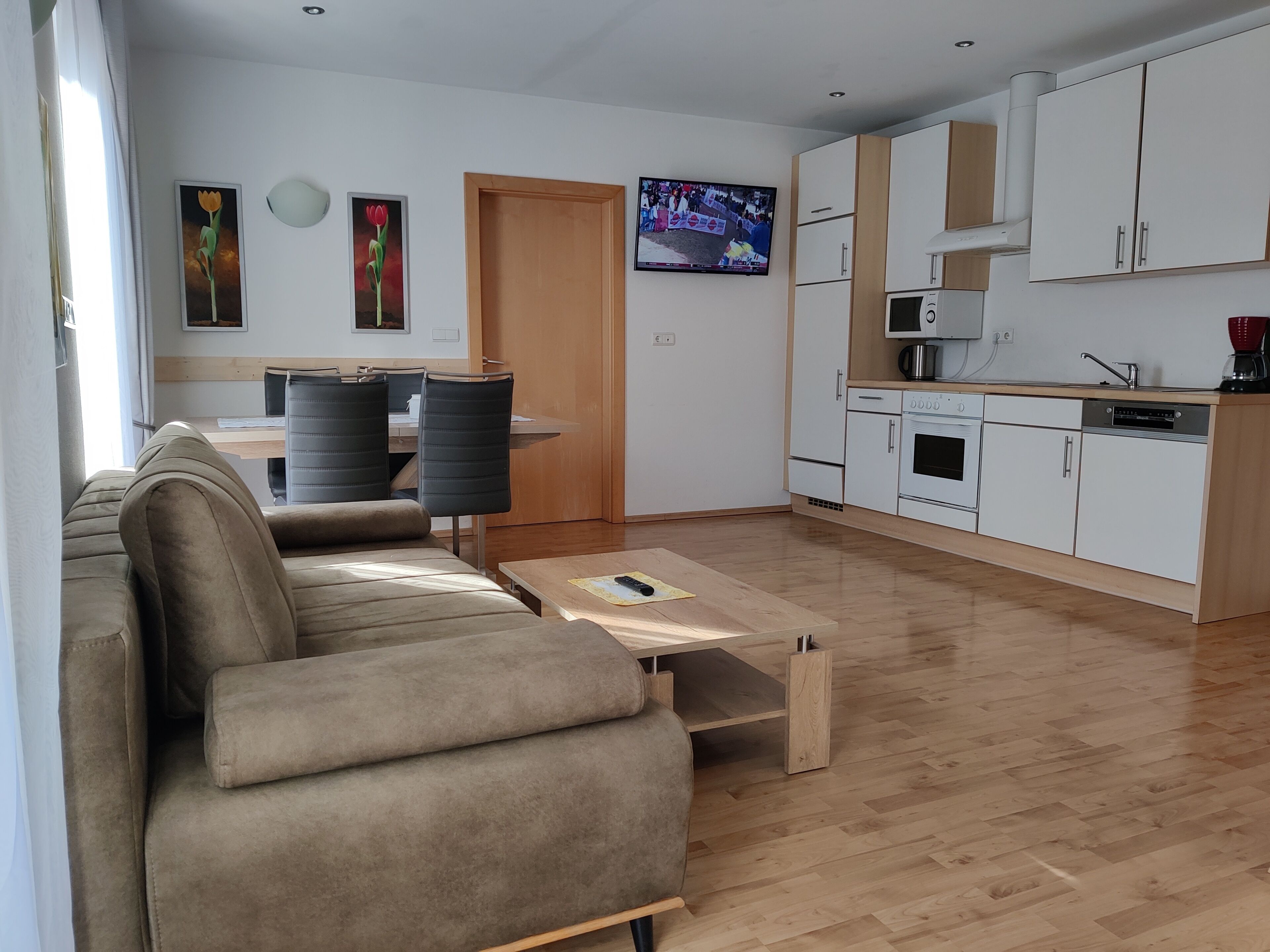 A modern living room with a sofa, a coffee table, and a dining area with chairs. There is a kitchen with white cabinets and appliances.