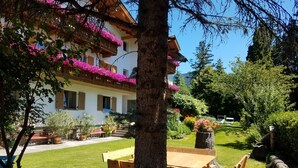 Un edificio con un balcone decorato da fiori rosa, un tavolo e panche in legno in primo piano, e un giardino curato con diverse piante e un albero grande.