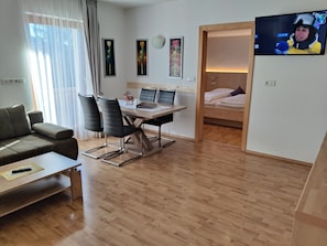A modern hotel room with a wooden floor, a dining area with a table and chairs, a sofa, a TV, and a bed with a wooden frame.