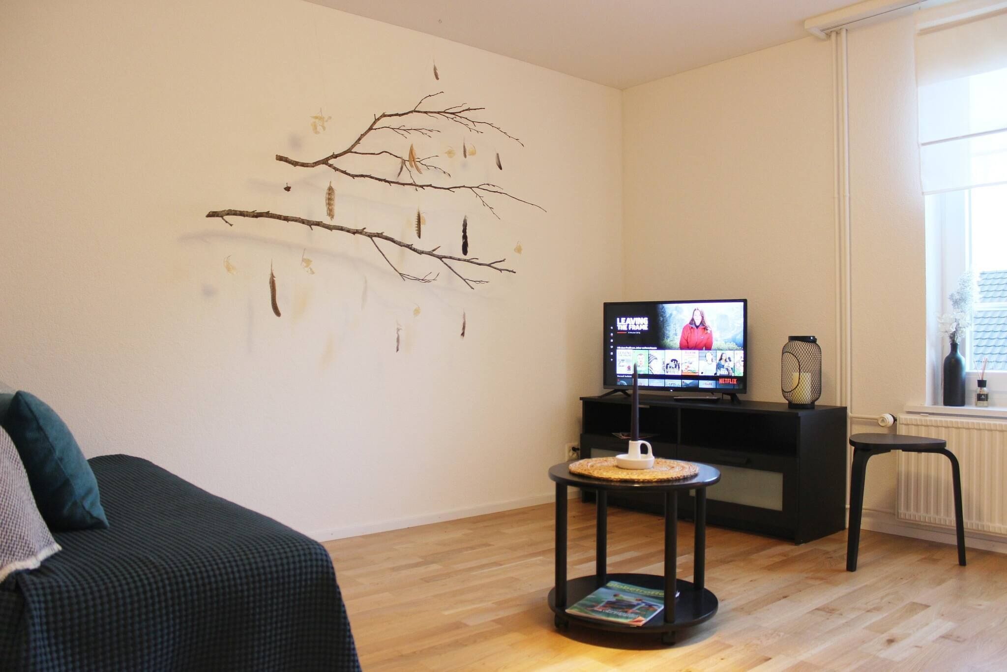 Ein Wohnzimmer mit einem Flachbildfernseher, Wandkunst aus einem dekorativen Zweig, einem kleinen runden Tisch mit einer Zeitschrift und einem schwarzen Sofa.
