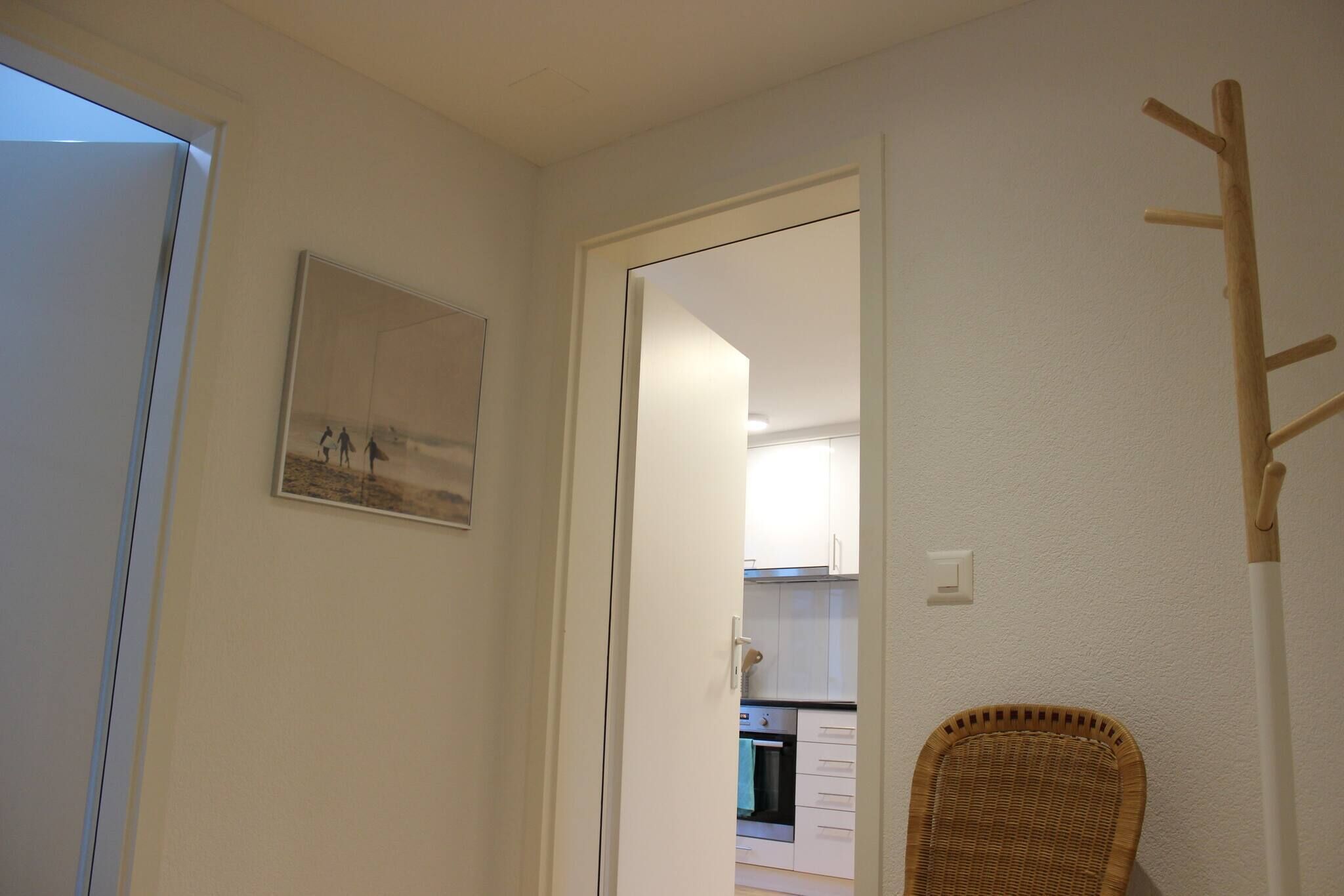 A hallway with a wicker chair, a door leading to a kitchen, and a framed picture on the wall.