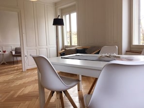 A dining area with a white table, grey chairs, a black floor lamp, and a view of the outdoors through large windows. - Casa Schilling - 4 Charming Rooms in the Best Location (St. Gallen)