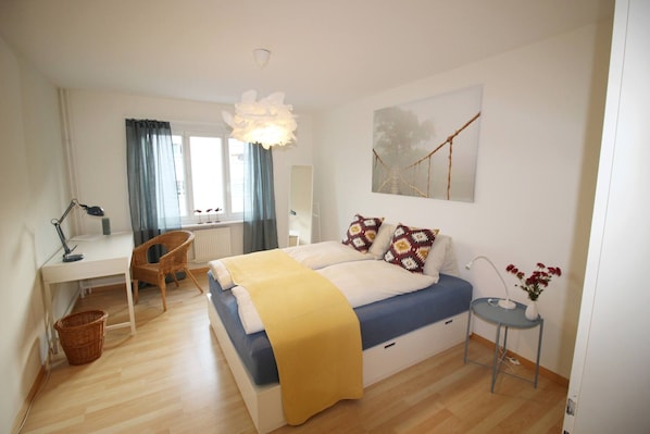 A bedroom with a bed, desk, chair, and a small table with a vase.