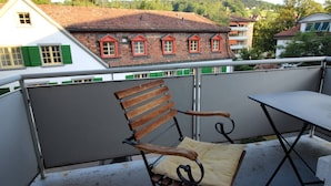 A balcony with a wooden chair and a table, overlooking a building with red window shutters and a tiled roof. - Casa Schilling- 2,5 Rooms With Balcony Near Hospital, University (St. Gallen)