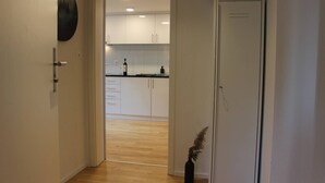 Un couloir mène à une cuisine dotée de meubles blancs et d’un plan de travail noir.