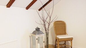 Une pièce avec un plancher en bois, une chaise en osier, une armoire blanche avec une porte en verre et un vase décoré de branches.