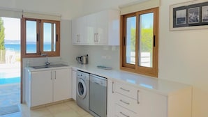Cocina moderna con armarios blancos, lavadora y vistas al mar a través de grandes ventanales.