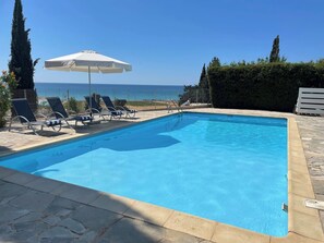 En klarblå pool med solstolar och ett parasoll, omgiven av stenplattor och med utsikt över havet.