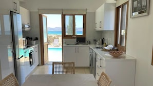 Una cocina moderna con armarios blancos, una mesa de comedor con sillas de mimbre y vistas al mar a través de amplias ventanas.
