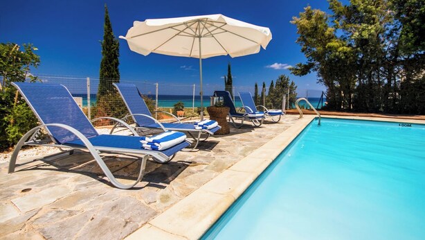 Zona de piscina con sillones de descanso y una sombrilla, con vista a un cielo azul despejado y al océano.