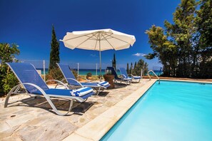 Un espace aménagé au bord de la piscine, avec des chaises longues et un parasol, offrant une vue sur un ciel bleu azur et l’océan.
