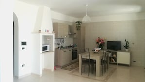 A modern kitchen with a microwave, stove, and hanging utensils. There is a dining area with a table and chairs, and a TV on a stand. - Bright Apartment a few km From the sea (Minervino di Lecce)