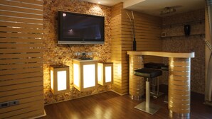 A modern living room with a flat-screen TV mounted on a brick wall, wooden slatted blinds, and a bar area with stools and a counter. - 2B - 2 Bedrooms and 2 Bathrooms Located Downtown Apartment With Shared Pool (Bangkok)
