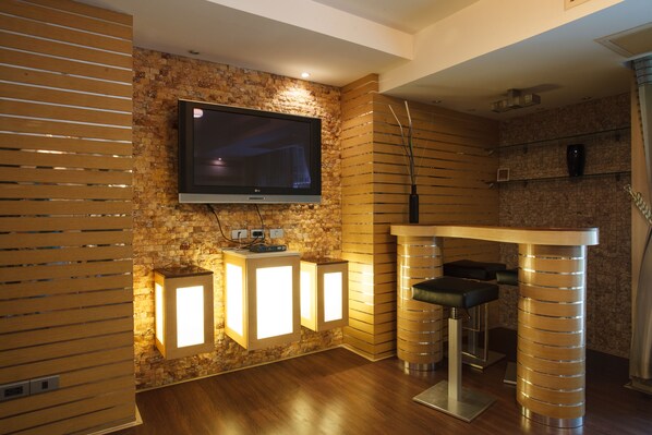 Una sala de estar moderna con un televisor de pantalla plana montado en una pared de ladrillo, persianas venecianas de madera y una zona de bar con taburetes y mostrador.