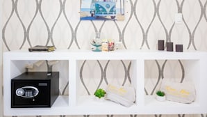 A wall with a geometric pattern, a framed picture of a van on a beach, a safe, and a shelf with towels and a plant.