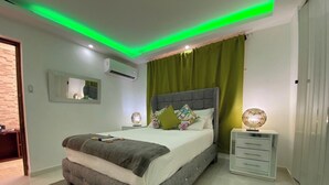 Une chambre à coucher moderne avec un lit recouvert d’un tissu gris, des rideaux verts, une lampe de chevet et une petite table de chevet blanche.
