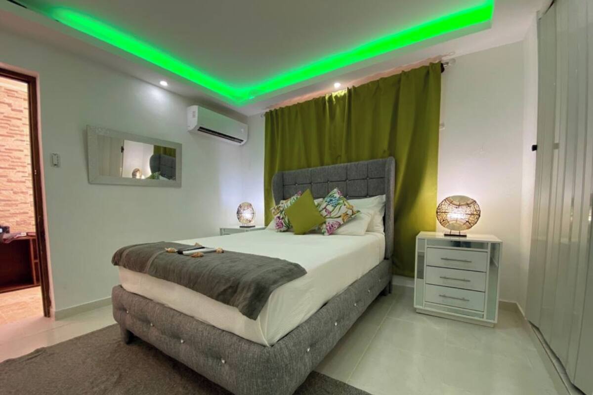 A modern bedroom with a grey upholstered bed, green curtains, a bedside lamp, and a small white nightstand.