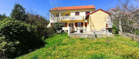 A two-story building with a red-tiled roof, a balcony, and a garden with yellow flowers.