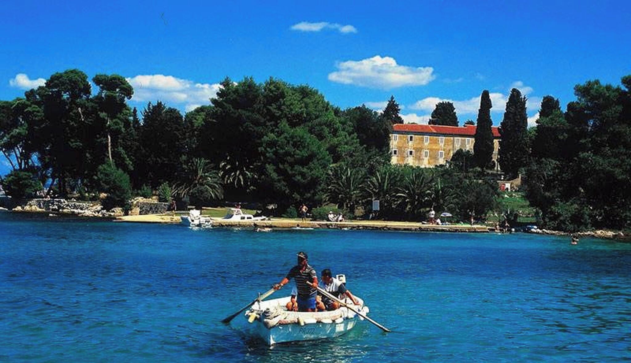 A boat on a clear blue sea with a building and trees in the background.