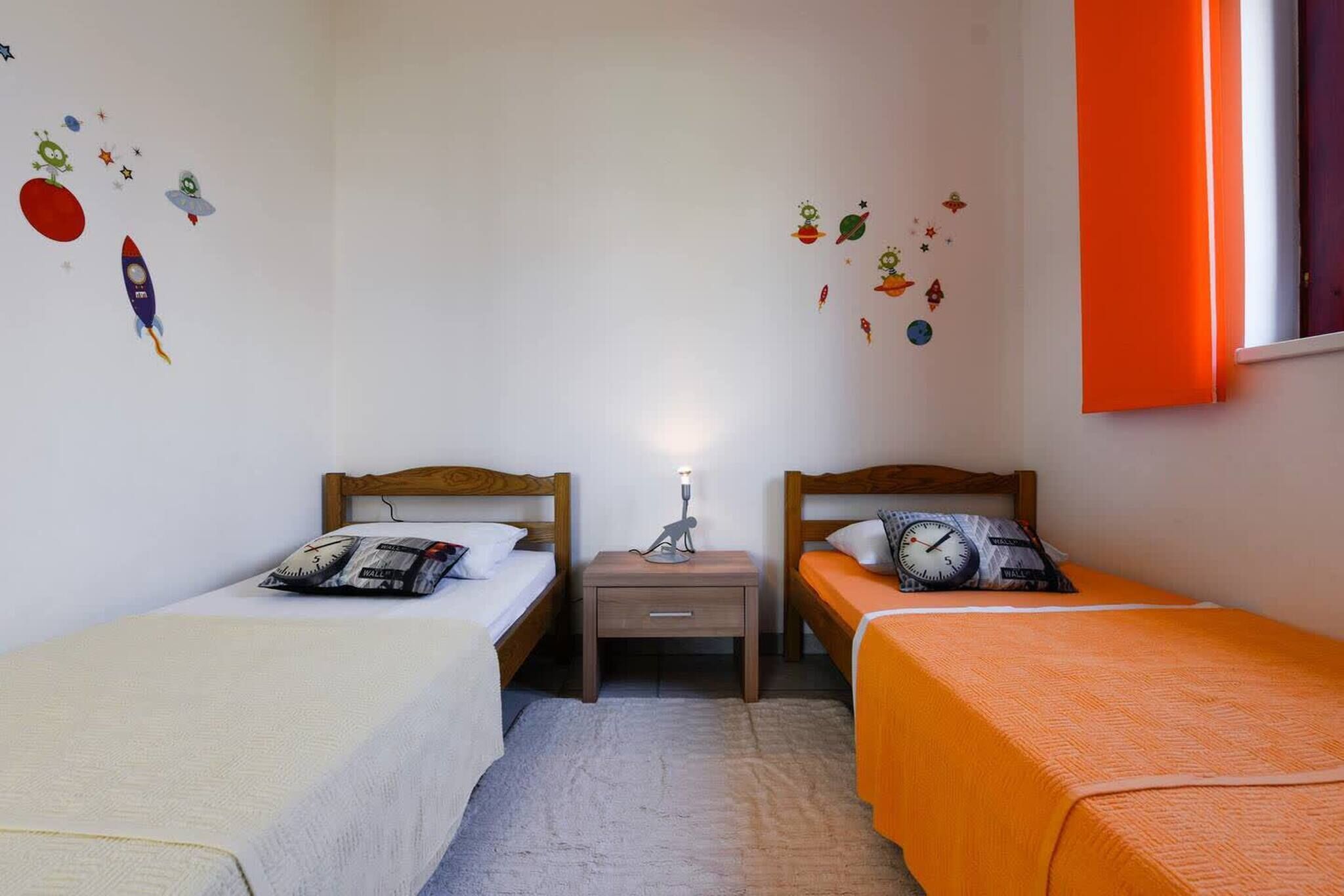 A room with two single beds, a nightstand with a lamp, and wall decals.