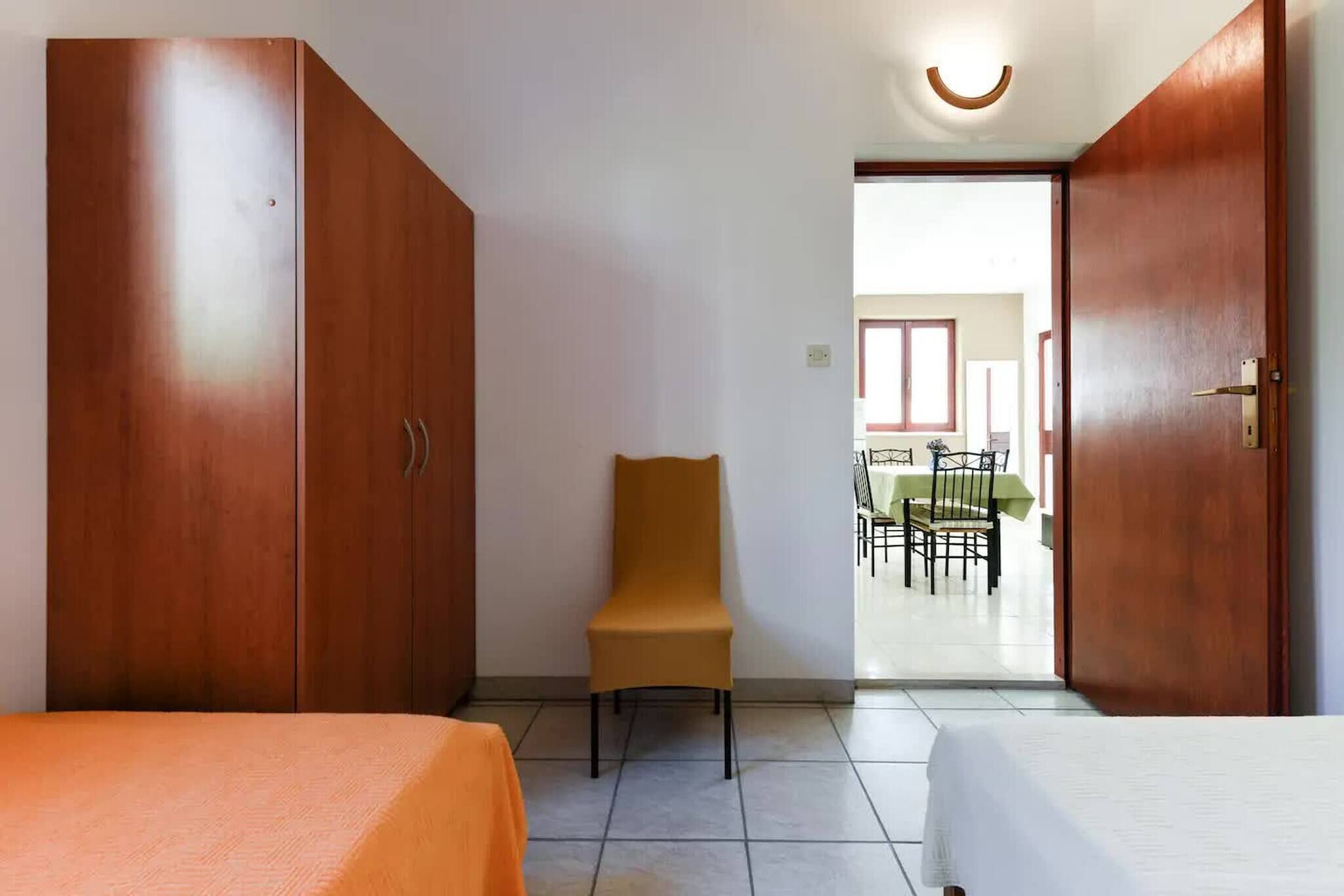 A room with a bed, a chair, and wooden wardrobes. There is a door leading to a dining area with a table and chairs.