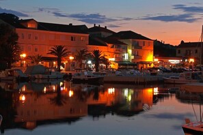 A marina with boats docked, buildings with illuminated signs, and palm trees.